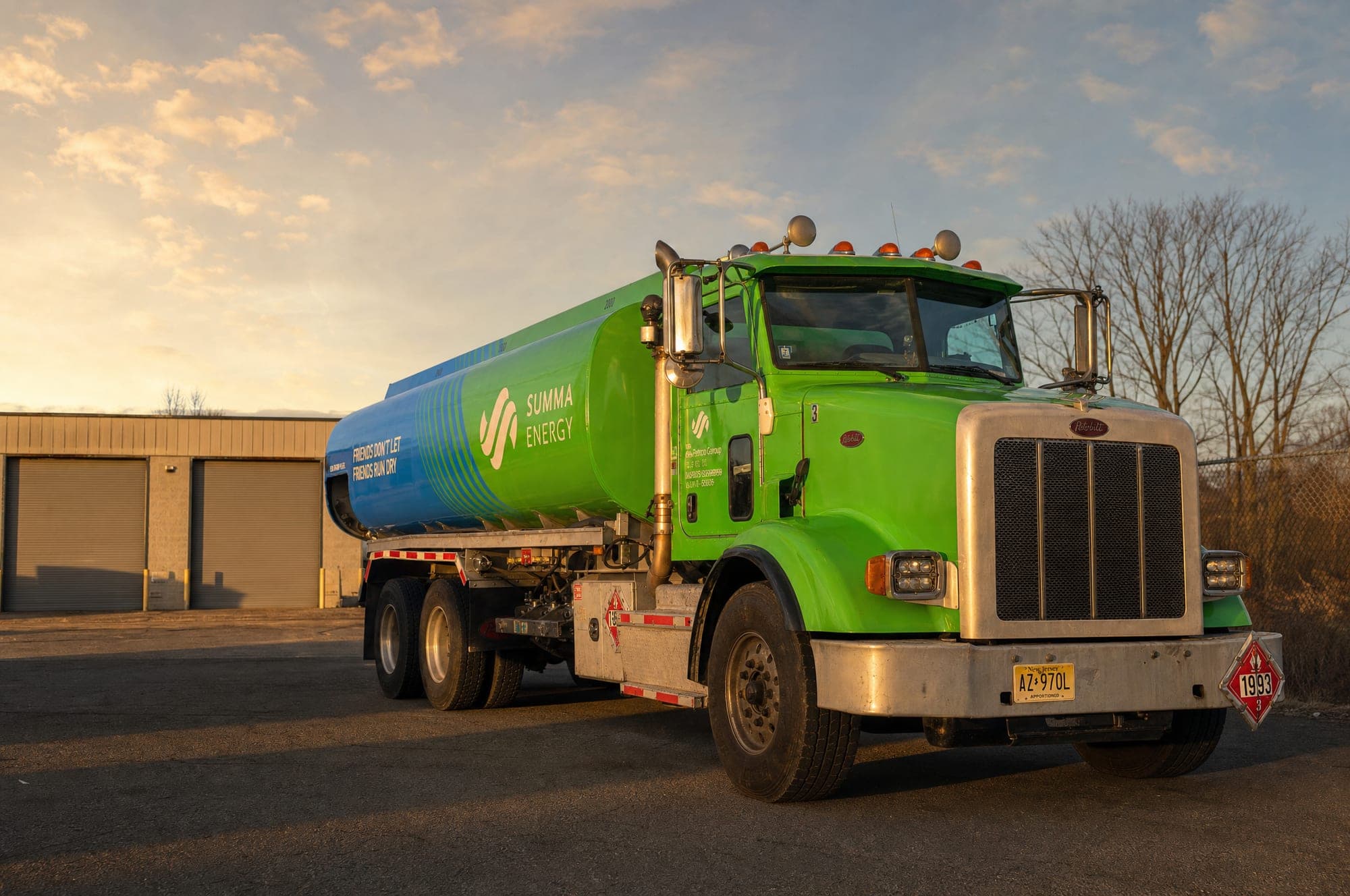A Summa Energy tanker parked on a depot apron in low winter sun, green cab and green-to-blue tank, seen from the front three-quarter with roller-shutter bays behind it.